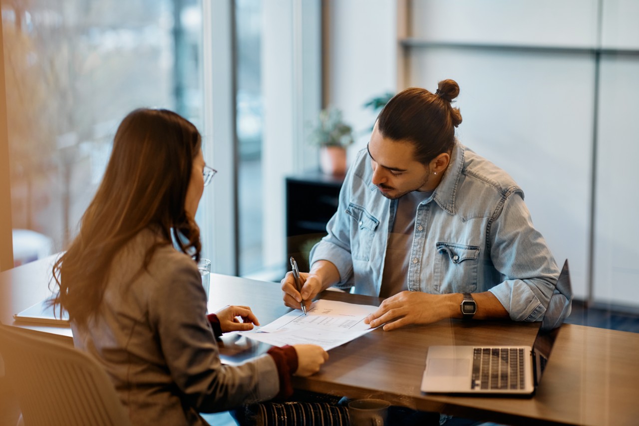 Young Hispanic man signing loan agreement during meeting with his bank manager in office. Young Hispanic man signing loan agreement during meeting with his bank manager in office.