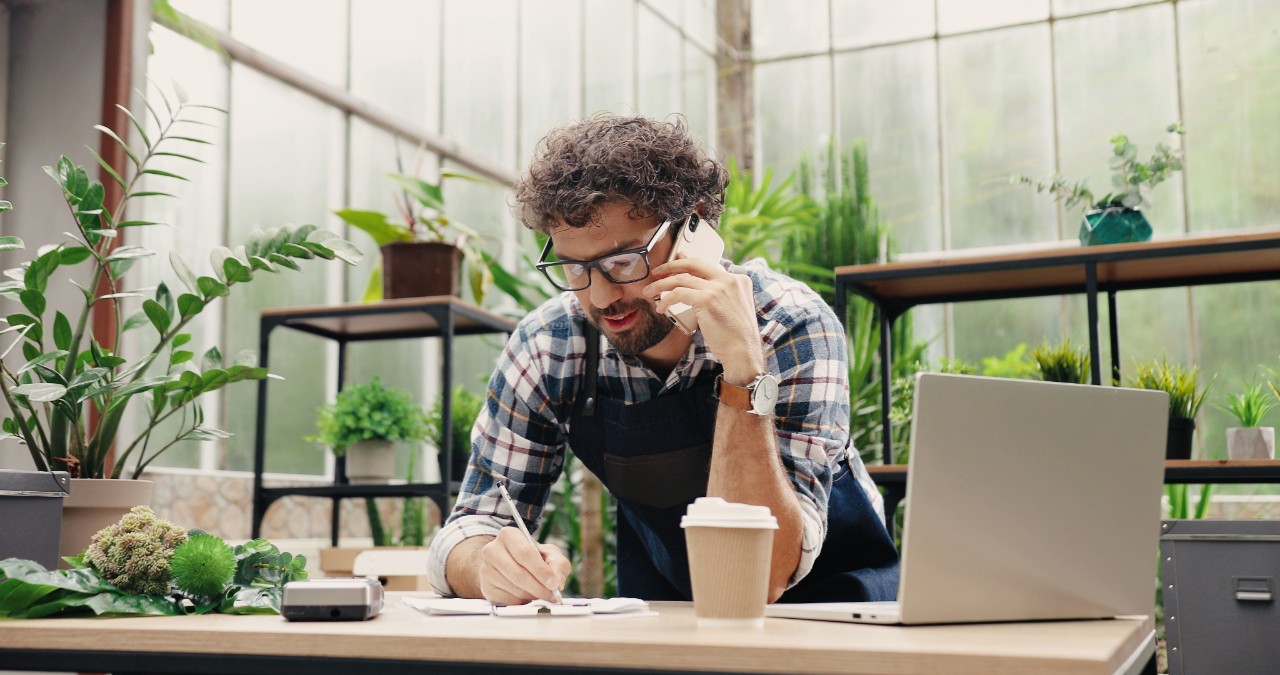 Happy Caucasian businessman talking on cellphone while standing in apron in small floral center and writing down order details. Joyful male florist calling on smartphone at work. Own business concept Fintech